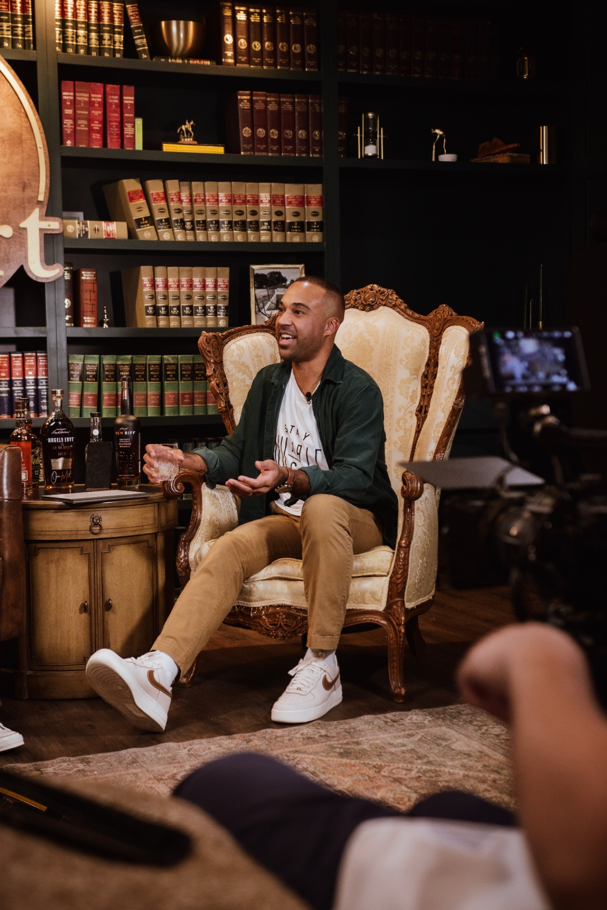 Arian Green raising a rocks glass mid-conversation, bourbon bottles staged in the foreground
