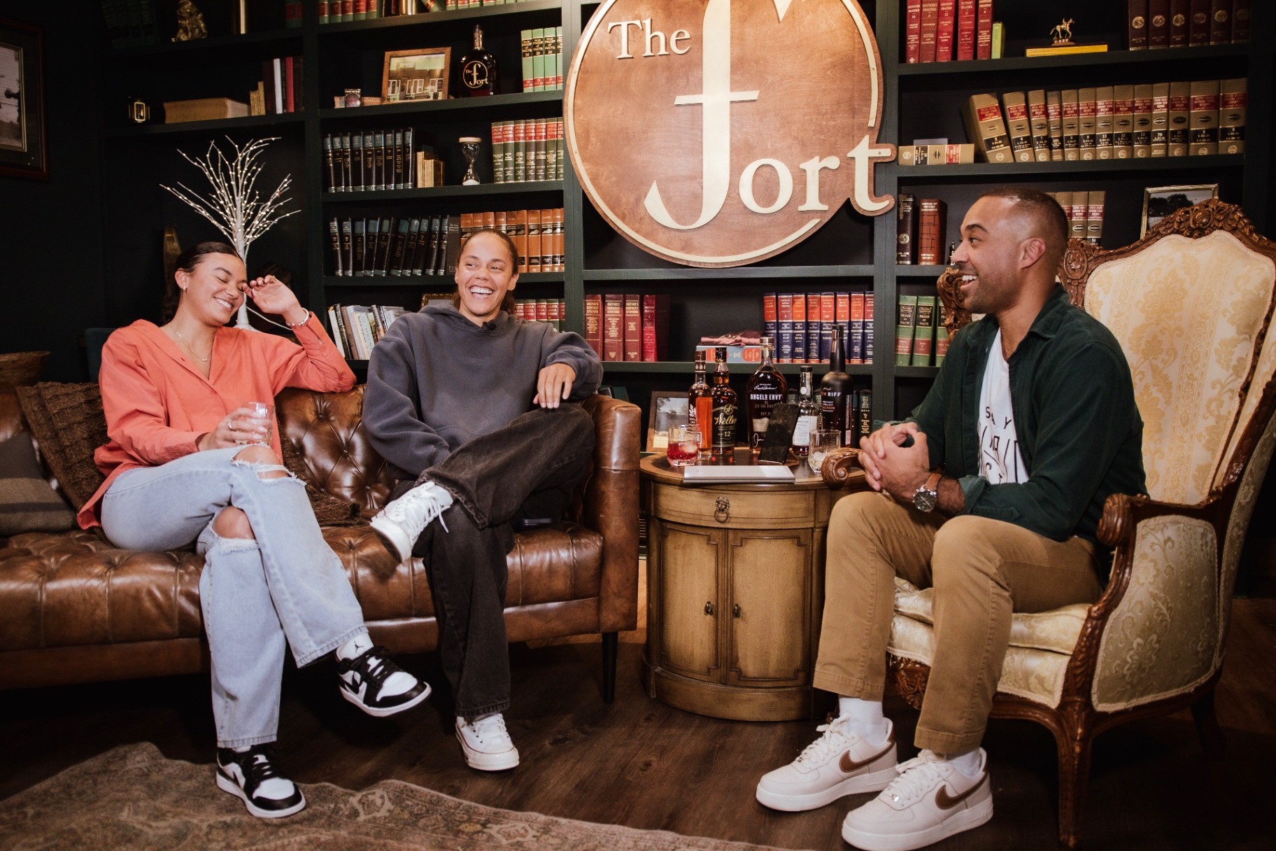 Arian Green sharing a laugh with two guests over a pour, the bourbon lineup between them beneath the copper 'The Fort' sign